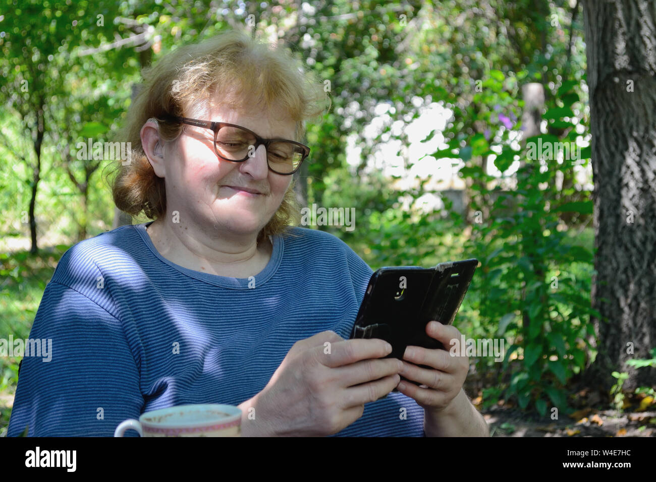 Frau mittleren Alters mit Brille im Garten sitzen und Lachen beim Browsen durch Social Media Netzwerke auf Ihrem Telefon. Stockfoto