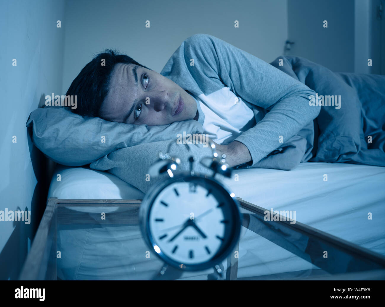 Schlaflose und verzweifelten jungen kaukasischen Mann wach nachts nicht schlafen zu können, frustriert und beunruhigt, schaut auf die Uhr leiden unter Schlaflosigkeit. Stockfoto