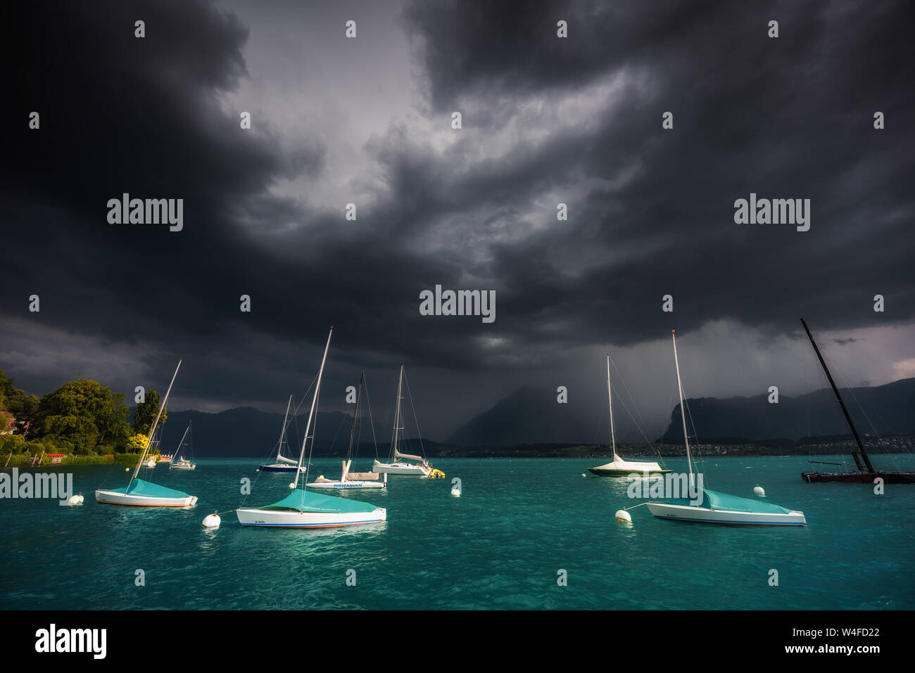 Sonnenbeschienene Boote der Segelschule Thunersee/Segelschule Thunersee mit einem dunklen Wetter vorne über die Berner Alpen, dunkle Wolken und Regen Stockfoto