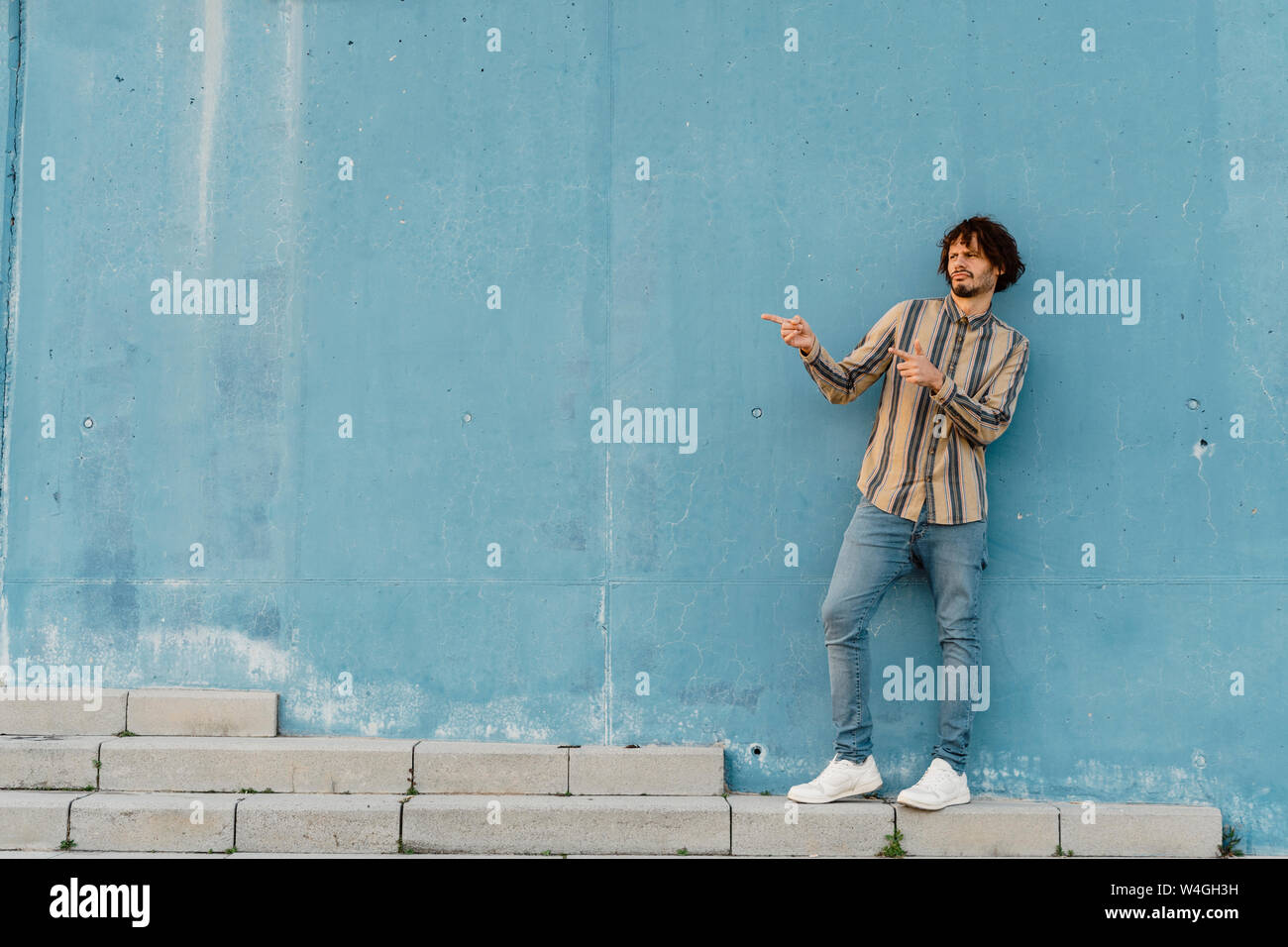 Der bärtige Mann mit gestreiften Hemd ständigen Ibn vor hellblau Wand gestikulierend Stockfoto