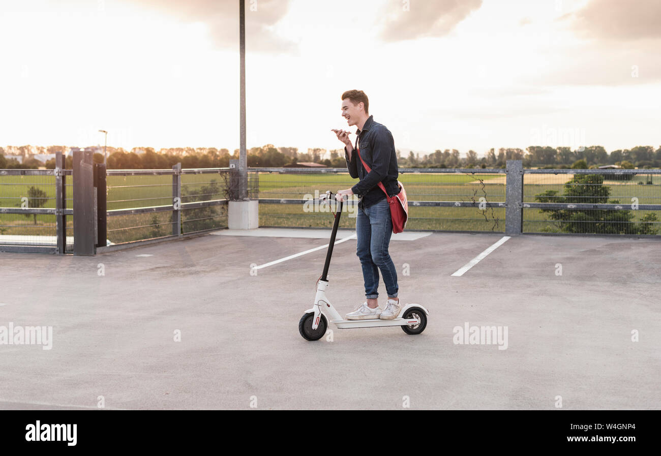 Glückliche junge Mann mit Handy und Elektroroller auf Parkdeck Stockfoto