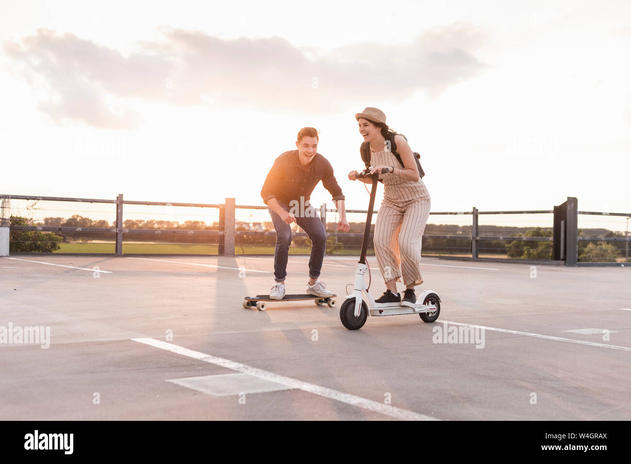 Junger Mann und eine Frau reiten auf Longboard und Elektroroller auf Parkdeck bei Sonnenuntergang Stockfoto