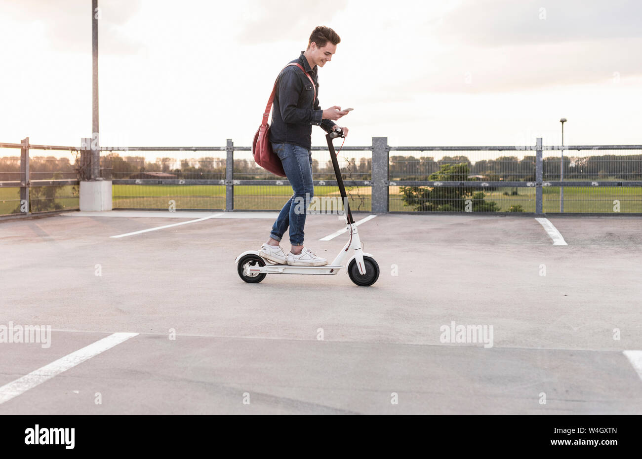 Glückliche junge Mann mit Handy und Elektroroller auf Parkdeck bei Sonnenuntergang Stockfoto