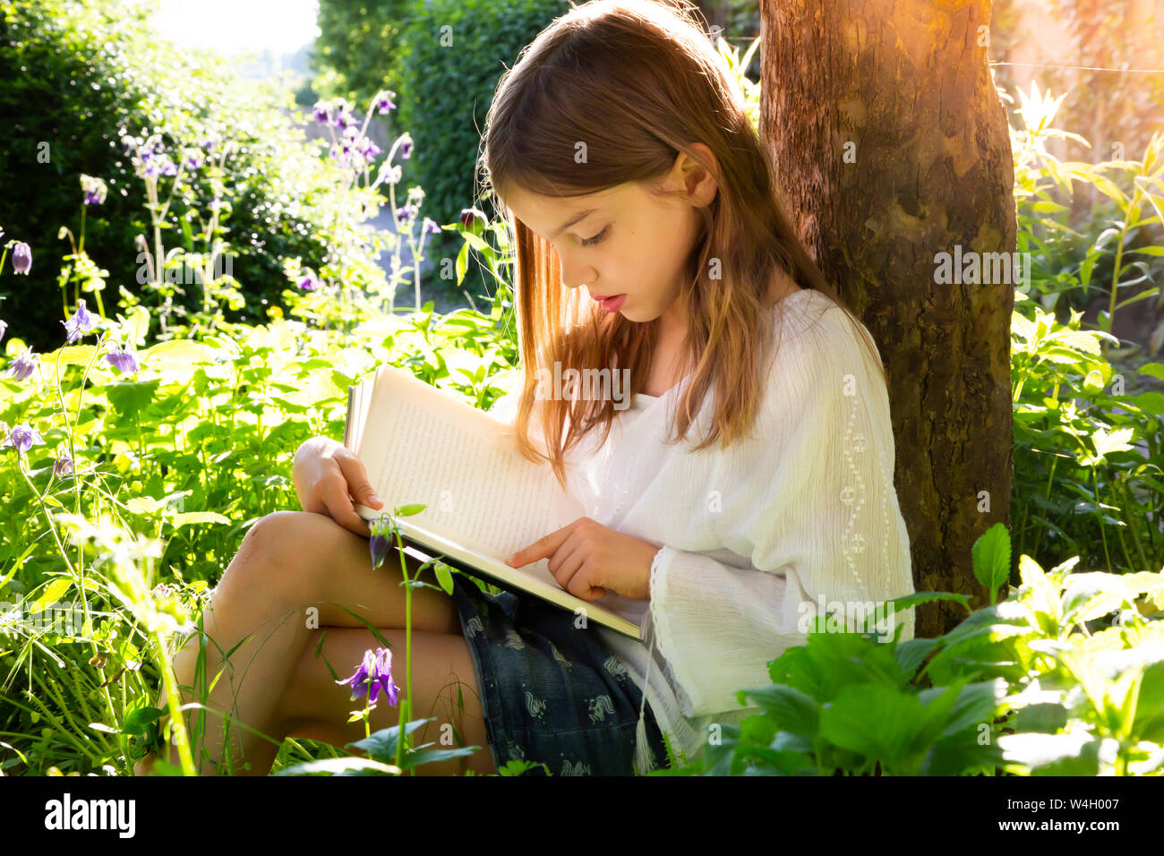 Kleines Mädchen gegen den Baumstamm gelehnt, ein Buch zu lesen Stockfoto