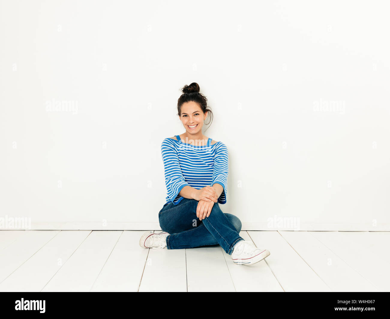 Schöne junge Frau mit schwarzen Haaren und blau-weiß gestreiften Pullover sitzen auf dem Boden vor weißem Hintergrund Stockfoto