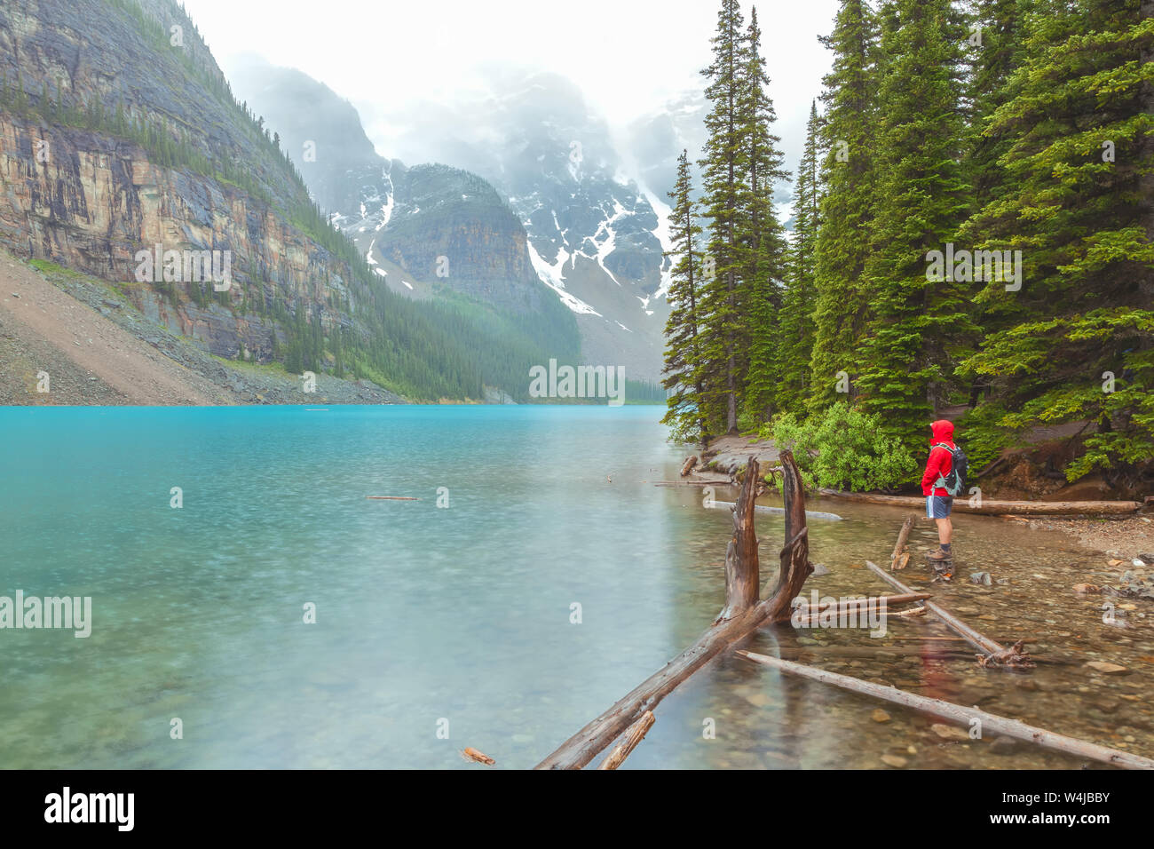 Mann in roter Regenjacke steht durch den Moraine Lake Shore unter dem Regen an einem Sommermorgen, Banff National Park, Alberta, Kanada. Stockfoto