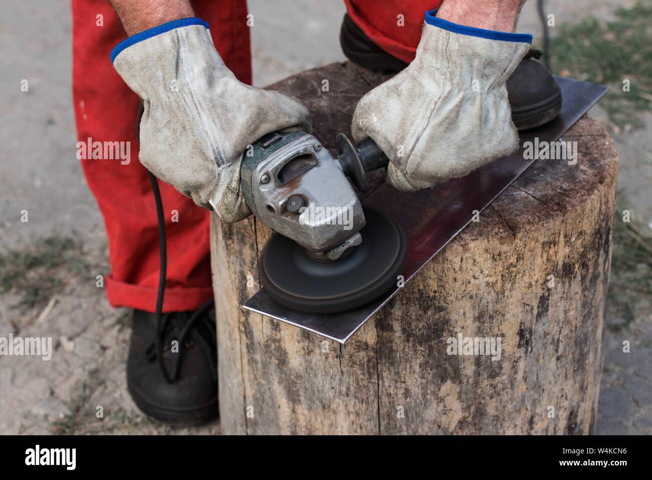 Der Master in einem roten Overall mit Händen in Arbeitshandschuhe schleift ein Stück Stahlblech mit einer alten Mühle Stockfoto