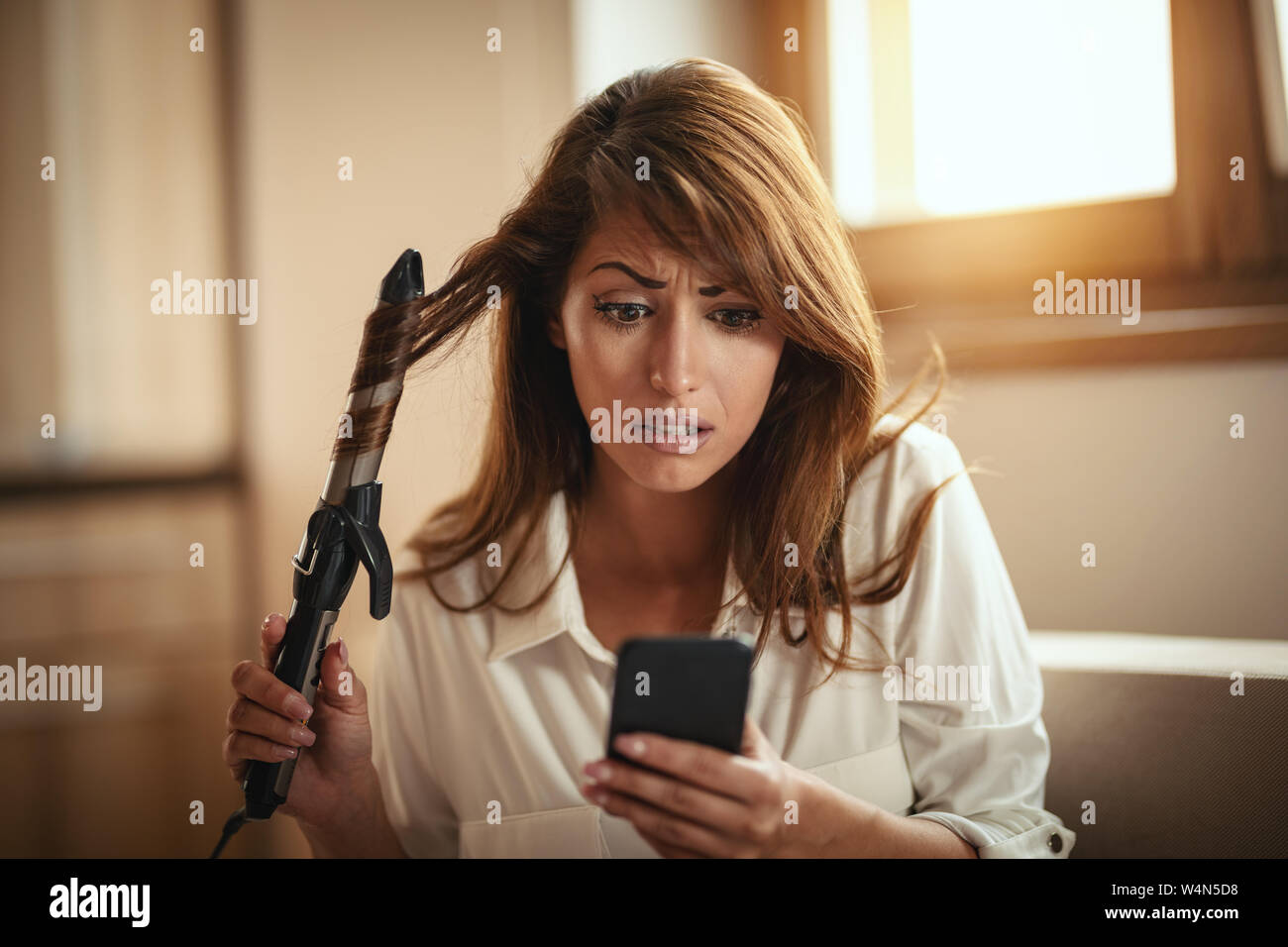 Eine junge Frau besorgt locken ihr Haar mit einem elektrischen Lockenwickler und schaut sich das Smartphone lesen Botschaft, die Sie nicht mögen. Stockfoto