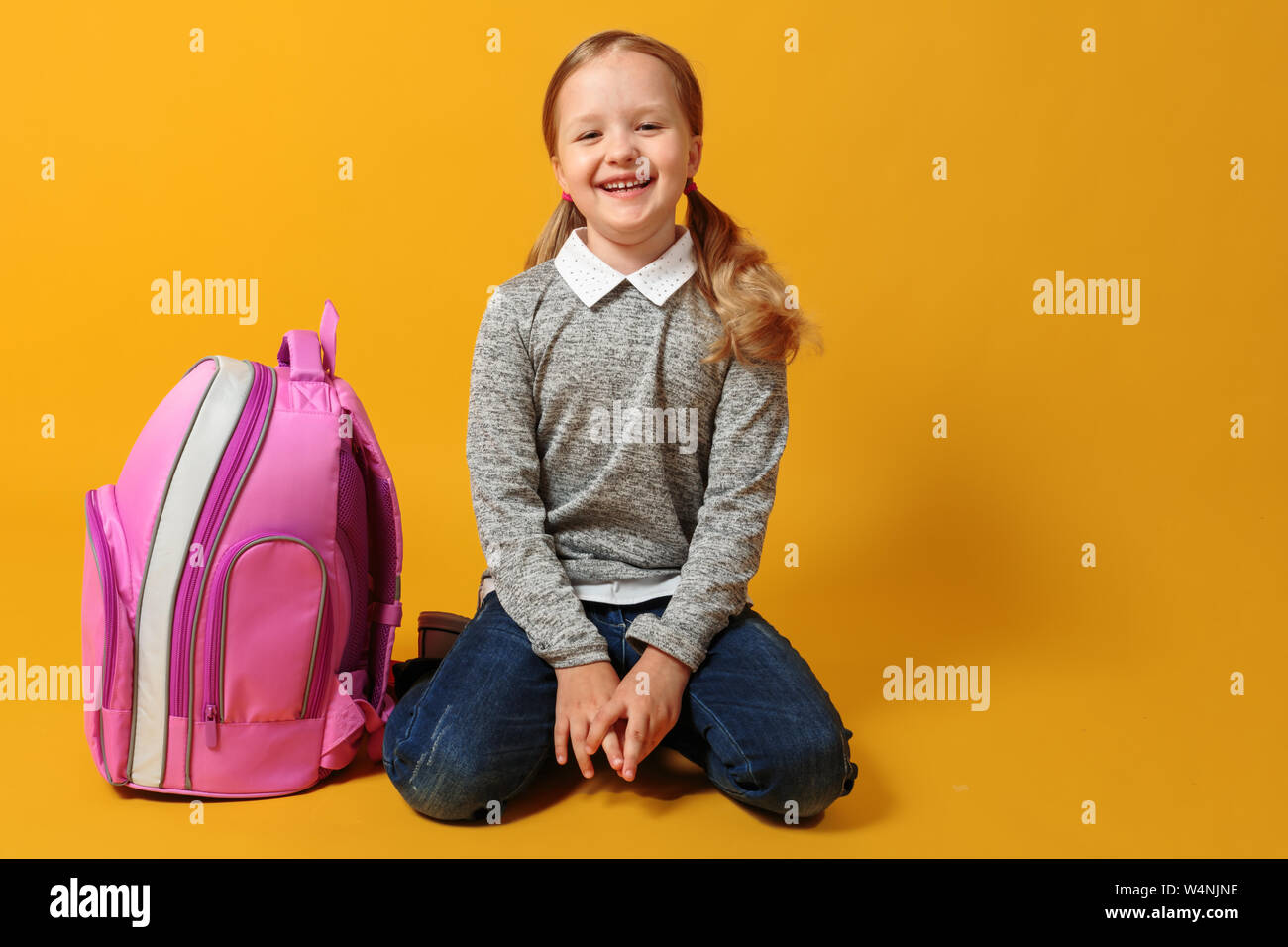 Fröhliche kleine Schüler Mädchen sitzt mit Rucksack auf dem Boden auf gelben Hintergrund. Der Begriff der Bildung. Zurück zu Schule. Stockfoto