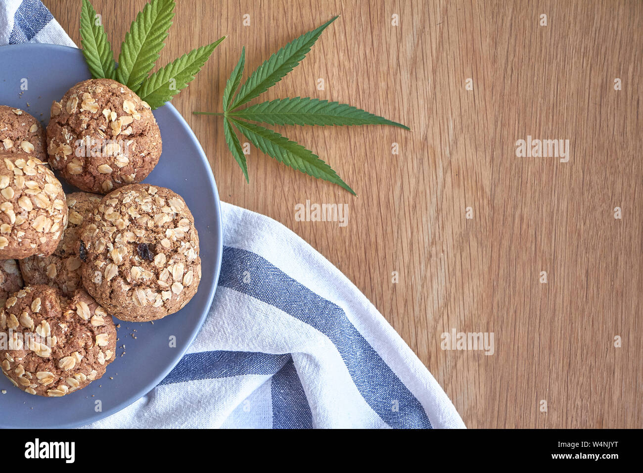 Ansicht von oben ein paar Haferflocken Topf Cookies auf graue Platte auf ein gestreiftes Handtuch auf hellem Holz- Oberfläche und zwei Blätter von Marihuana neben, selektiver Fokus Stockfoto