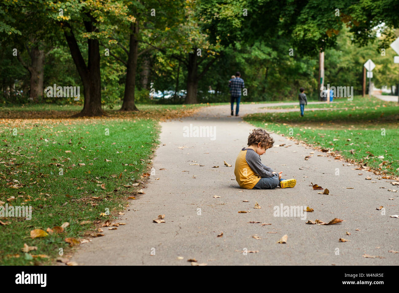 Ein kleiner Junge sitzt auf einem Blatt - übersäte Weg als seine Familie Spaziergänge auf Stockfoto