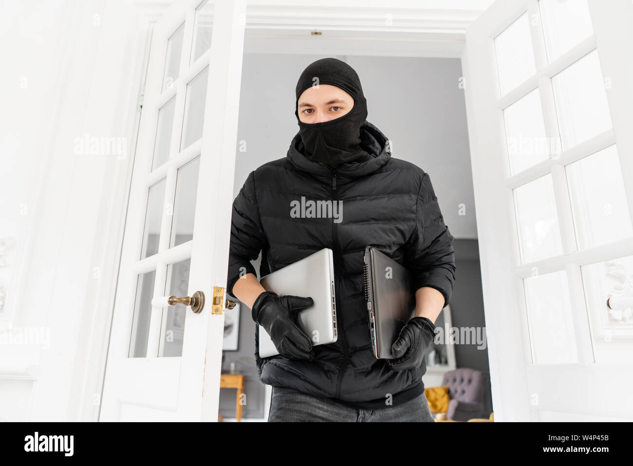 Dieb in die Kamera schaut mit schwarzer Sturmhaube stehlen zwei moderne teure Laptops. Der Einbrecher ein Verbrechen begeht in Luxus Wohnung mit Stuck. Stockfoto