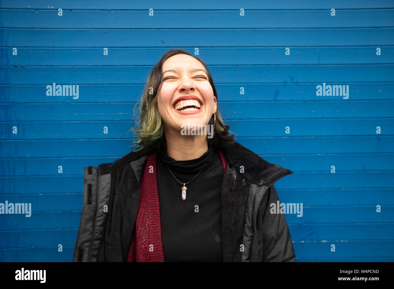 Lachende junge Frau mit geschlossenen Augen auf der blauen Wand, im Regen. Stockfoto