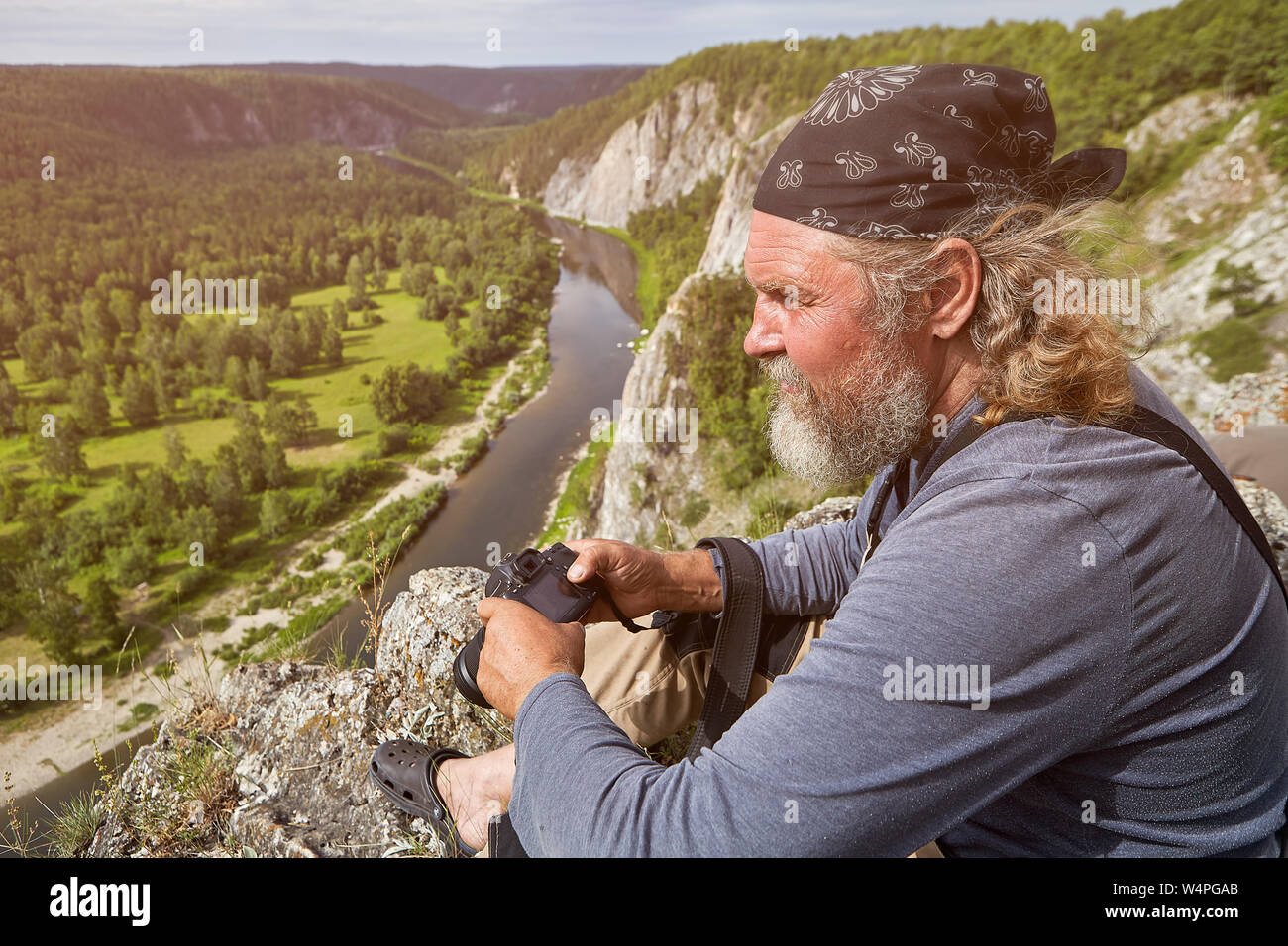 Ein reifer Mann mit einem Bart und in einem Bandana, auf seinem Kopf gebunden, liegt auf einer Klippe mit einer Kamera in der Hand, darunter eine Waldlandschaft mit ist Stockfoto