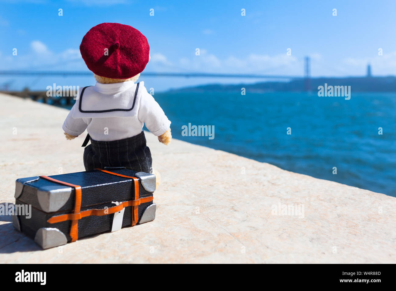 Wenig Teddybär verschleiß Matrosenanzug und barett Hut, auf Koffer im Quay in Lissabon, Portugal, Sitzen, Blick über Wasser zu überbrücken, träumen von Reisen Stockfoto