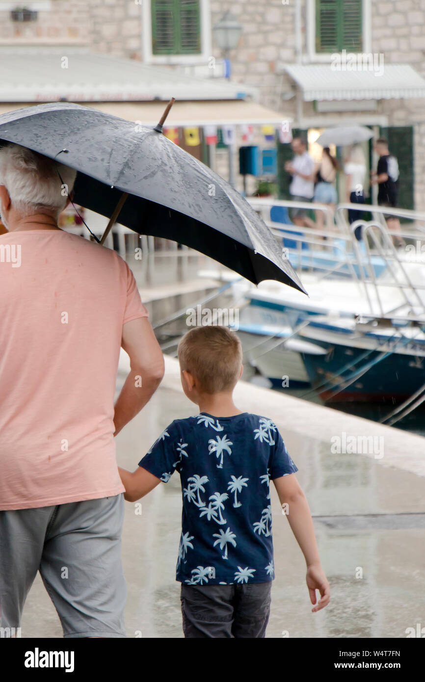 Vodice, Kroatien - 13. Juli 2019: Junge und sein Großvater gehen unter Dach in an einem regnerischen Sommertag am Meer Pier Stockfoto