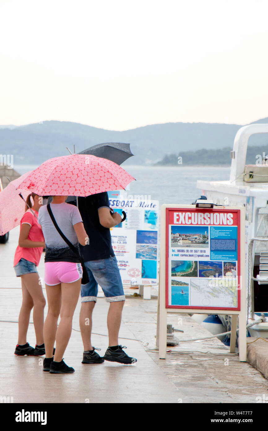 Vodice, Kroatien - Juli 13, 2019: Familie holding Sonnenschirme vor bootsausflug Werbetafeln an einem regnerischen Sommertag am Meer Stockfoto