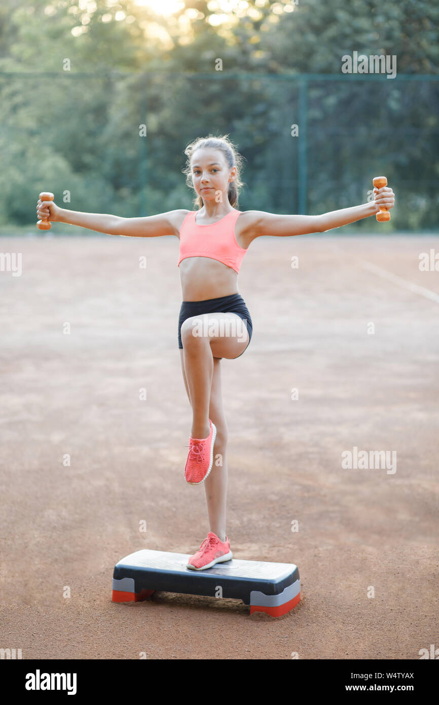 Ein nettes, hübsches junges Mädchen hält Hanteln in die Arme und führt verschiedene Übungen im Freien. Lebensstil Stockfoto