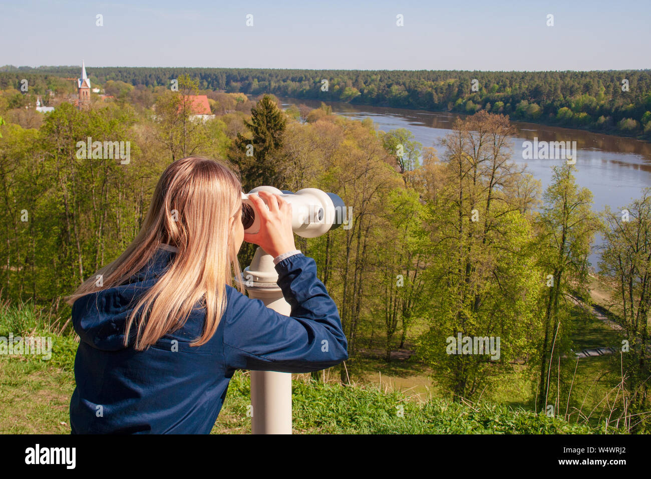Junge blonde Frau durch ein Fernglas, Wald, Fluss und die Dächer der Stadt auf der Suche nach Hintergrund Stockfoto