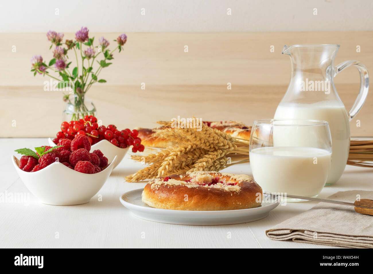 Weißer Tisch mit Yeasty süßes Gebäck mit frischen Johannisbeeren und Himbeeren und Streusel Topping, Glas und Krug Milch, garbe von Weizen, kleines Glas Stockfoto
