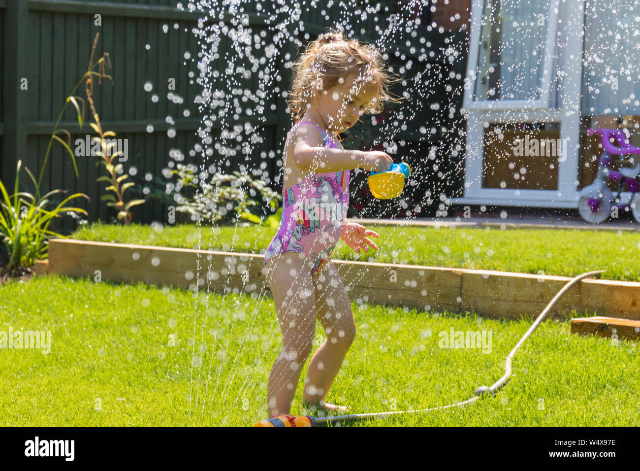 Glückliches, kleines Kind, das während der Sommerhitze 2019 unter einem Gartenwasserregner kühl spielt. England, Großbritannien spielerisch lernen Stockfoto