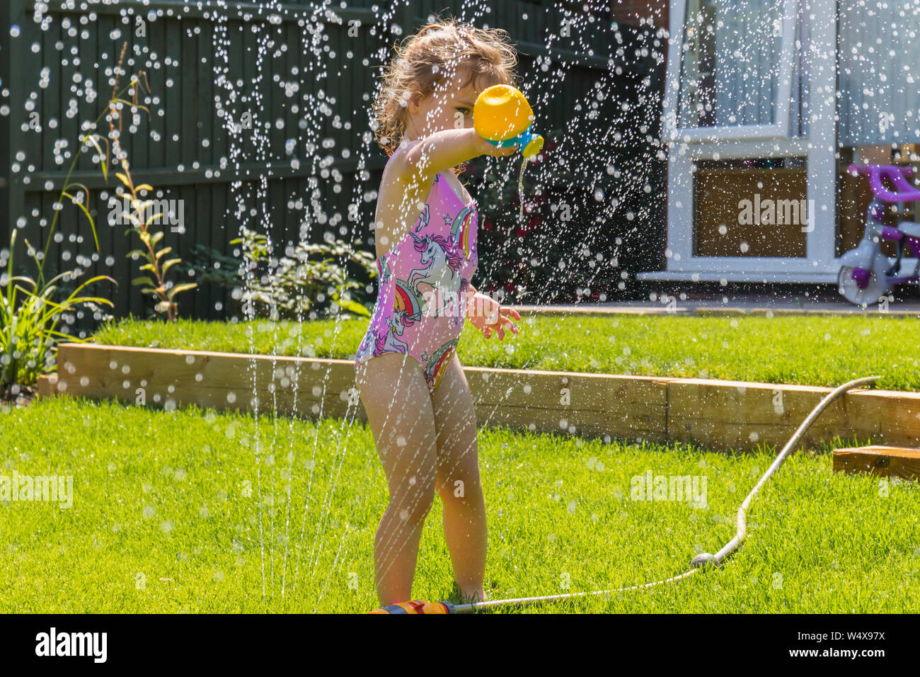 Glückliches, kleines Kind, das während der Sommerhitze 2019 unter einem Gartenwasserregner kühl spielt. England, Großbritannien spielerisch lernen Stockfoto
