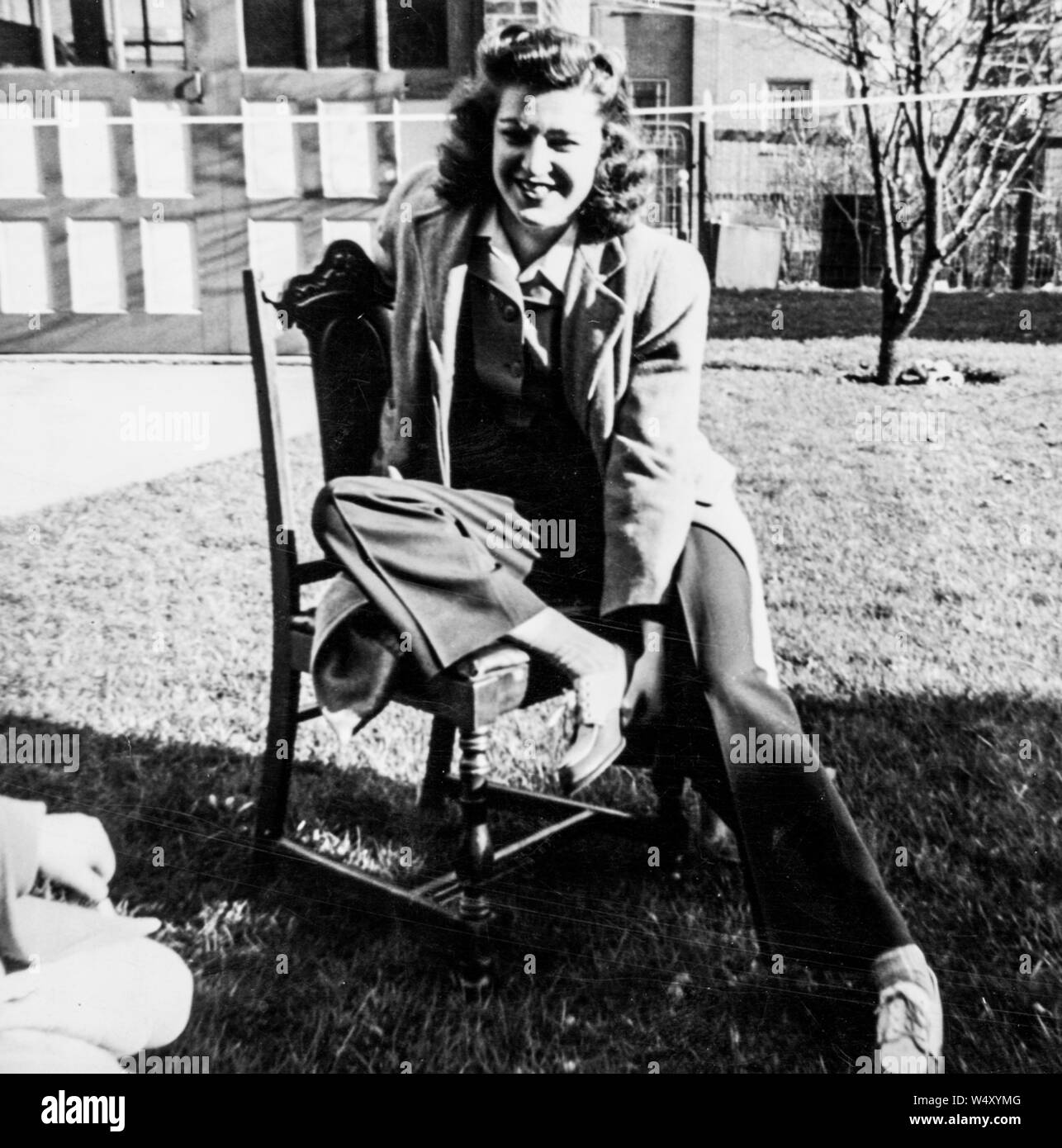 In voller Länge Porträt der jungen Frau sitzt auf einem Stuhl im Freien im Halbschatten, mit einem Bein auf den Stuhl und trug einen Mantel und Hose und lächelnd, 1950. () Stockfoto