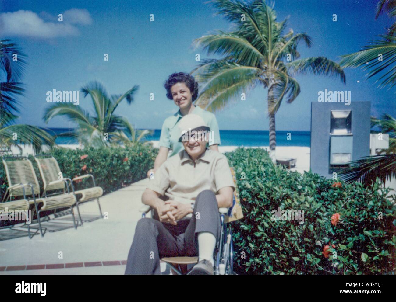 Reifen amerikanischen Touristen Paar in einer Lage am Strand in der Karibik, mit Mann im Rollstuhl sitzt, Brille und Hut, während seine Frau hinter ihm steht das Tragen von Lime Green Kleid, beide lächeln, 1970. () Stockfoto