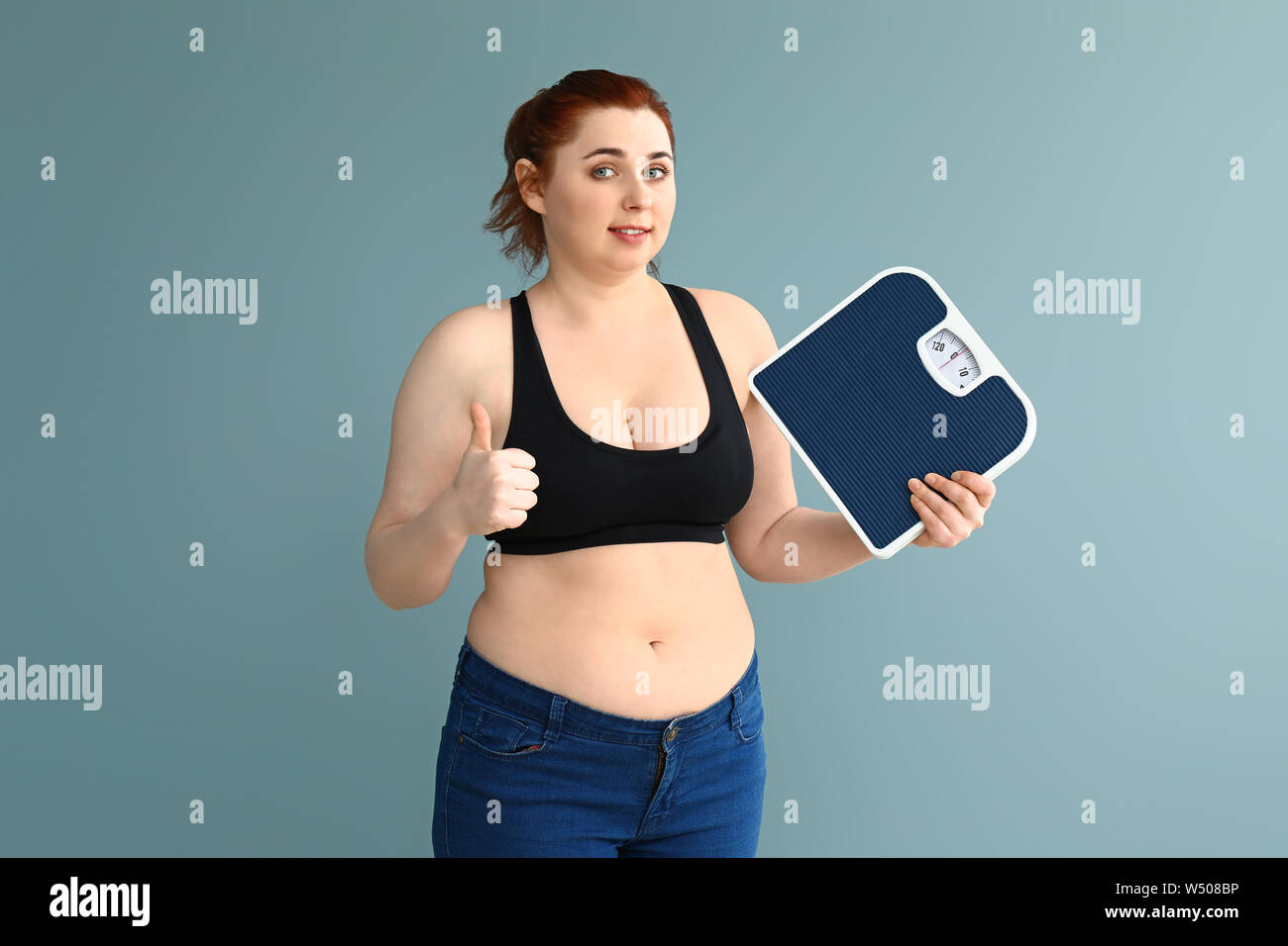 Übergewichtige Frau mit Skalen mit Daumen auf farbigen Hintergrund. Gewicht-verlust-Konzept Stockfoto