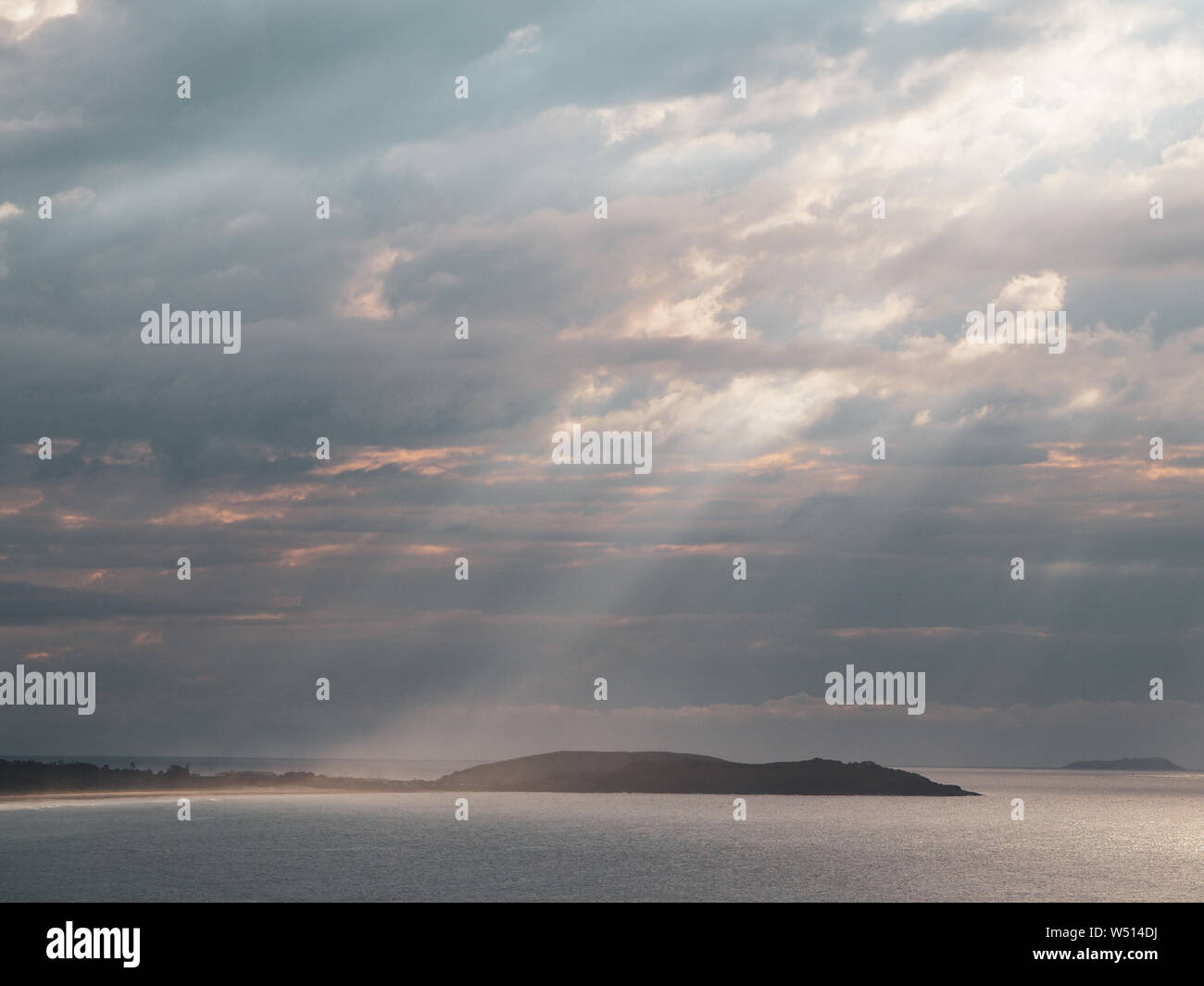 Sonnenlicht, das durch blaugraue Sturmwolken bricht, Strahlen, die ein rosa Leuchten auf Meer, Strand, schimmerndes Meer und Seascape-Szene strahlen Stockfoto