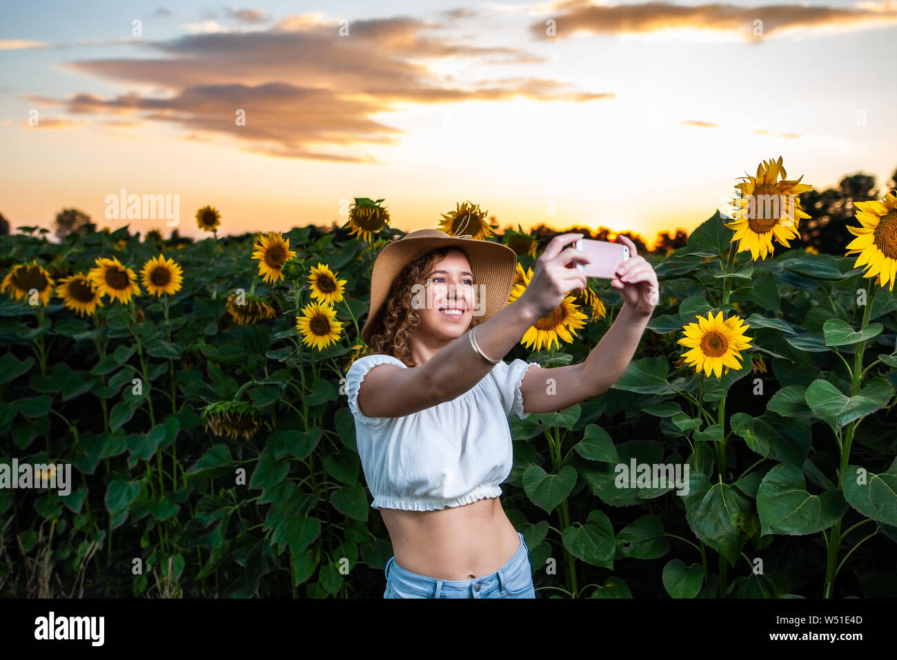 Schöne lockige Haare Frau Modell einer selfie mit Ihrem Smartphone vor einer Sonnenblume Wiese bei Sonnenuntergang Stockfoto