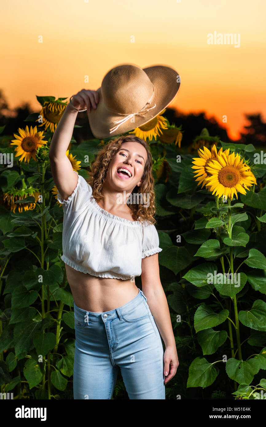 Schöne lockige Haare Frau Modell schwingen Ihren Hut vor einer Sonnenblume Wiese bei Sonnenuntergang Stockfoto