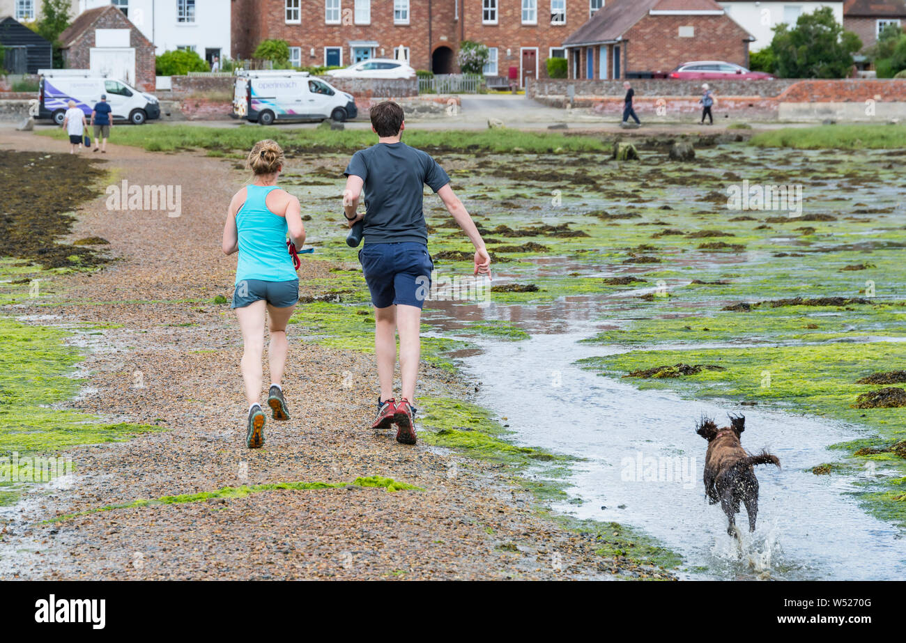 Ein junges Paar, das mit einem Hund über Bosham Causeway bei Ebbe in Chichester Harbour, Bosham Village, West Sussex, England, UK. Stockfoto