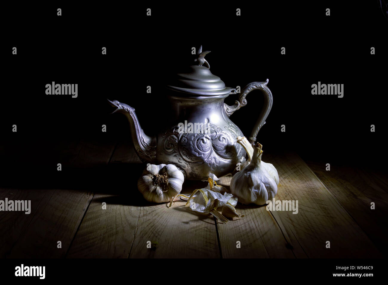 Stillleben mit dunklem Hintergrund. Eine orientalische Metall Teekanne im Gegenlicht, stehend auf einem Holztisch mit Knoblauch. Stockfoto