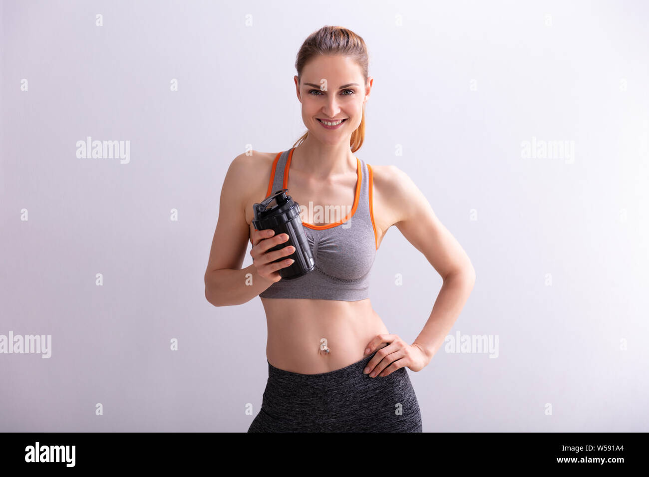 Portrait von lächelnden Fitness Woman Holding Protein Shaker Flasche gegen grauer Hintergrund Stockfoto