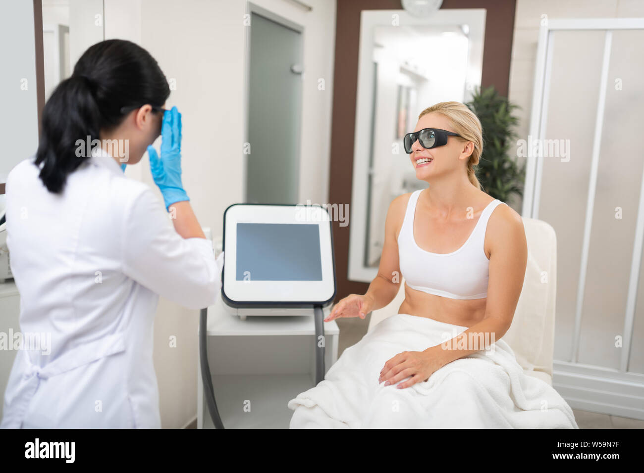 Halten schön. Lächelnde Frau sitzen in Schutz brille in einem Schönheitssalon und Beginn ihrer Verfahren mit der Kosmetikerin. Stockfoto