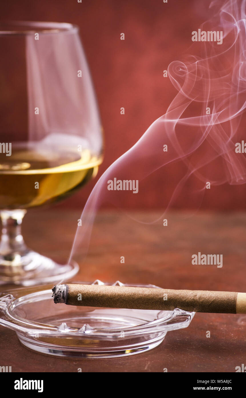 Leuchtet eine Zigarre mit Rauch Aschenbecher im Vordergrund, im Hintergrund ein Glas mit Alkohol Stockfoto