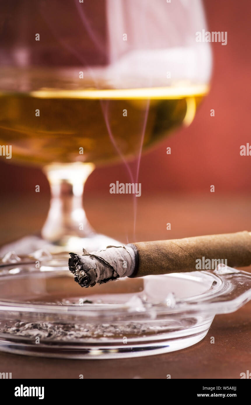 Leuchtet eine Zigarre mit Rauch Aschenbecher im Vordergrund, im Hintergrund ein Glas mit Alkohol Stockfoto