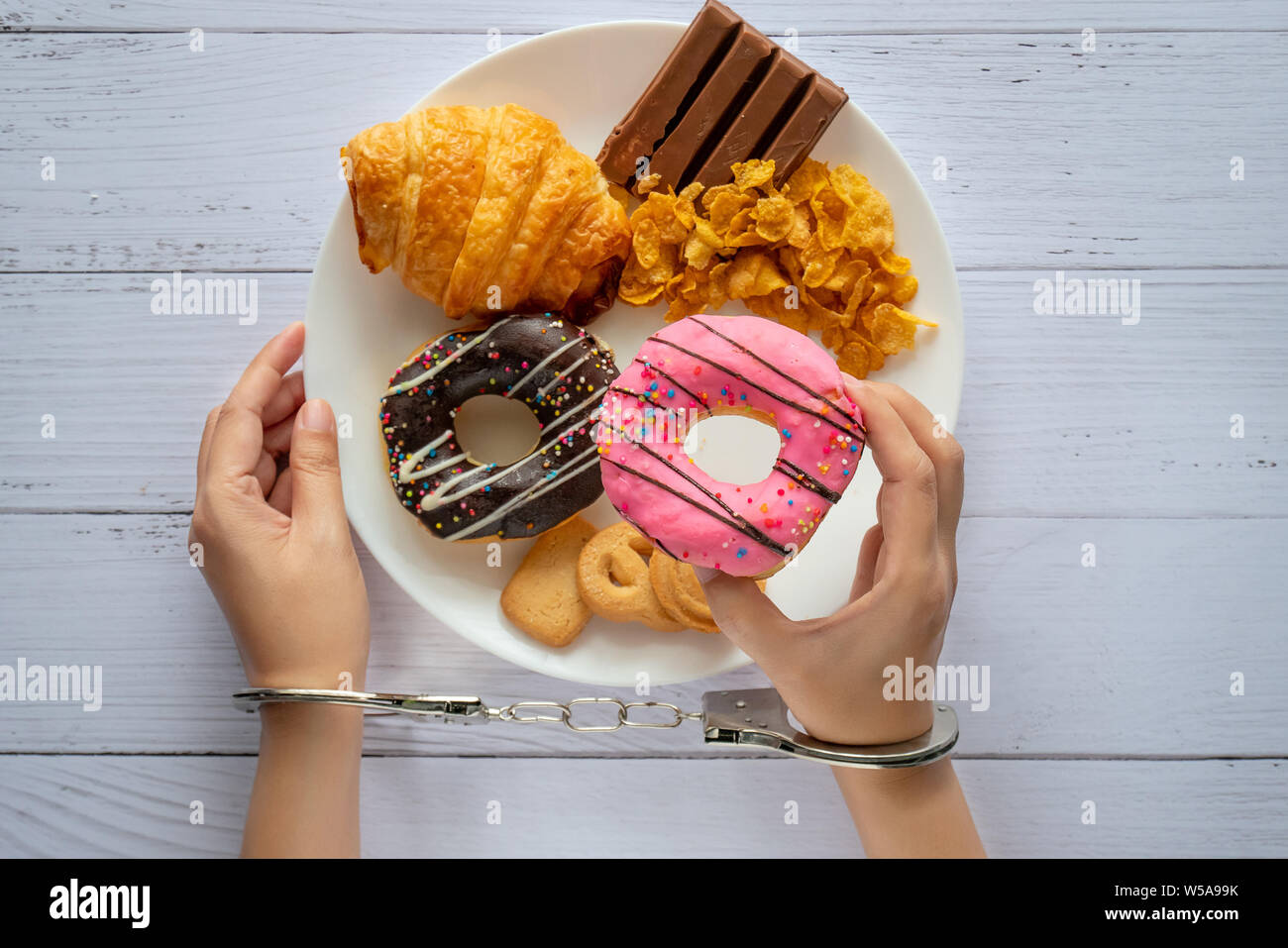 Kalorien, Ernährung und Gewichtsabnahme Konzept. nach oben Blick auf zwei Händen war Kontrolle durch Handschellen, holding Donut aus weißen Teller auf hölzernen Tisch Stockfoto