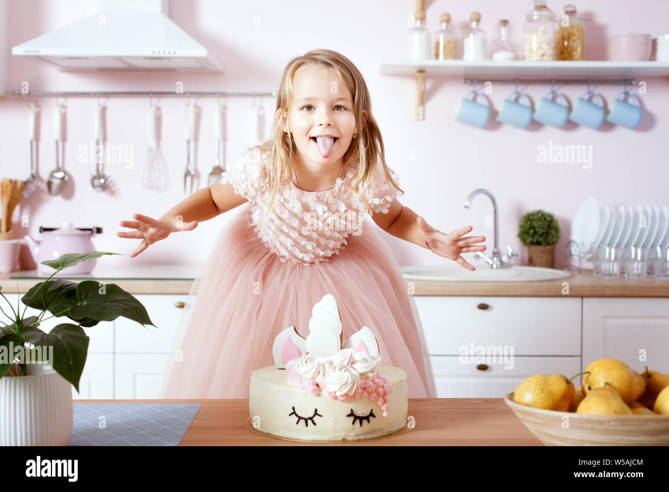 Kleines Mädchen steht in der Nähe der Tabelle mit einem Geburtstagskuchen und zeigt Zunge. Stockfoto