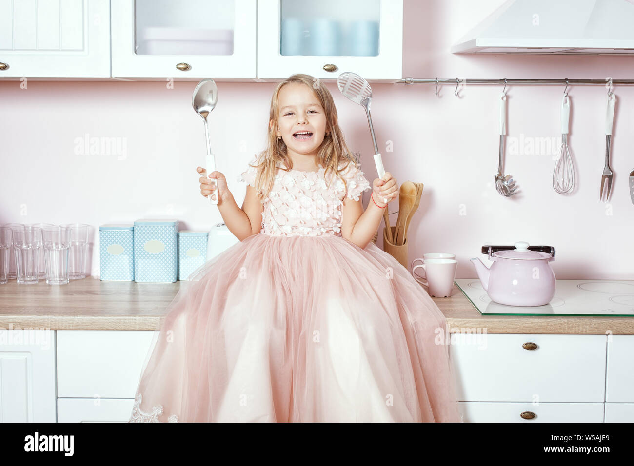 Wunderschöne kleine Mädchen, das in einem prächtigen Kleid sitzt auf dem Tisch in der Küche und lacht. Stockfoto
