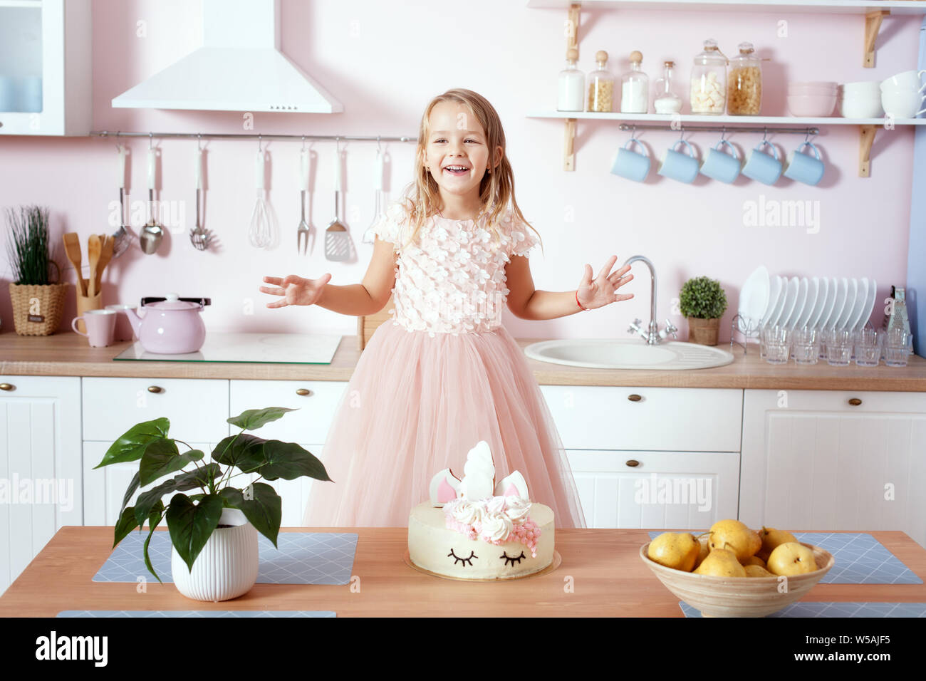 Kleines Mädchen in einem schönen Kleid steht in der Küche an den Tisch. Auf dem Tisch ist eine Geburtstagstorte. Stockfoto