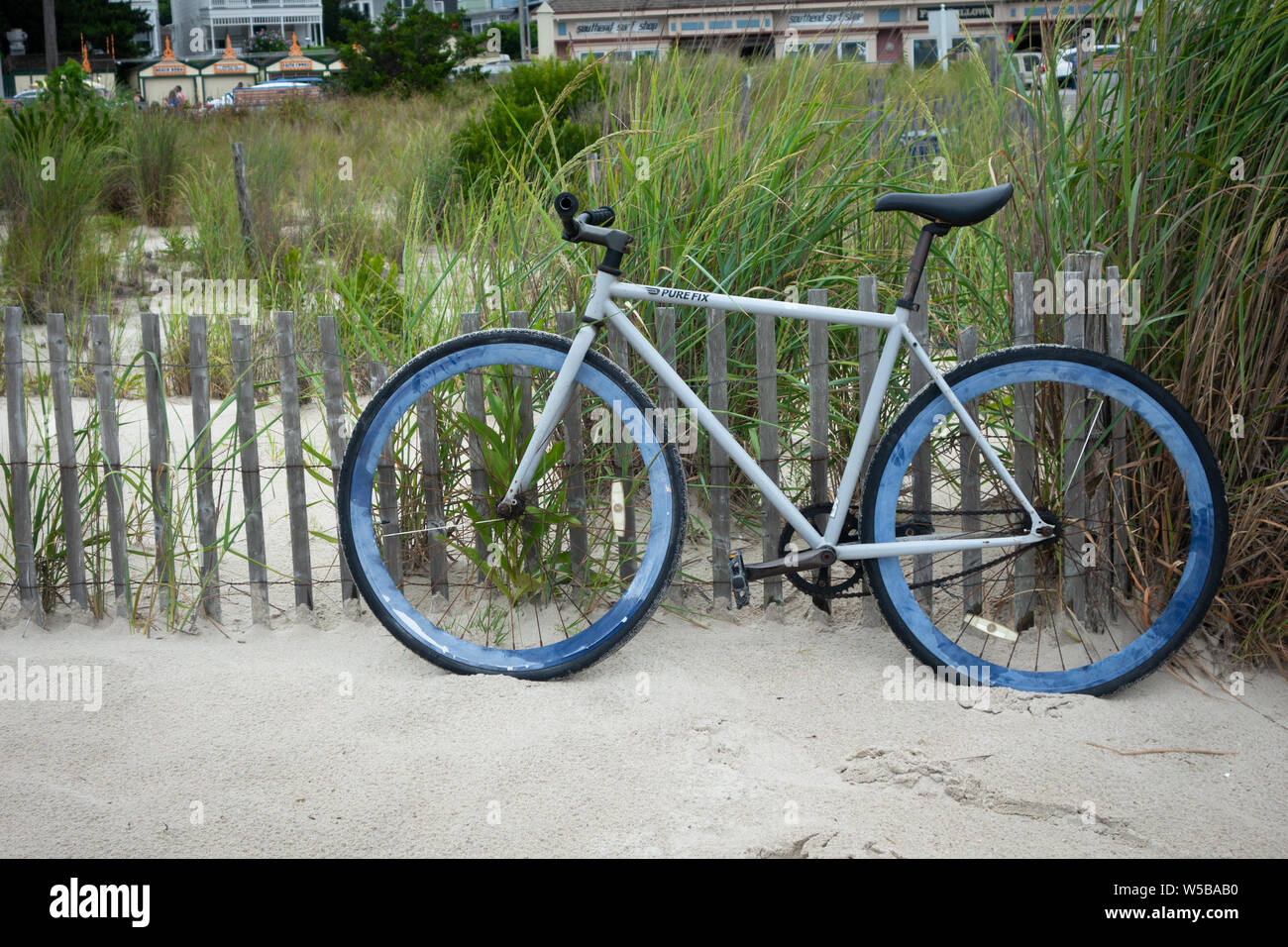 Geparkte Fahrräder, die an einen Holzzaun auf einem Sandstrand gelehnt sind, umgeben von hohem Gras und weit entfernten Gebäuden Stockfoto