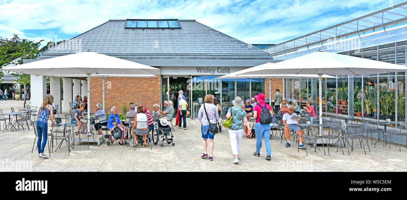 RHS Wisley Leute gehen zu neuen, modernen Cafe Gebäude Eingang mit Sonnenschirm Schatten & outdoor Tische Royal Horticultural Society Gärten Surrey England Großbritannien Stockfoto