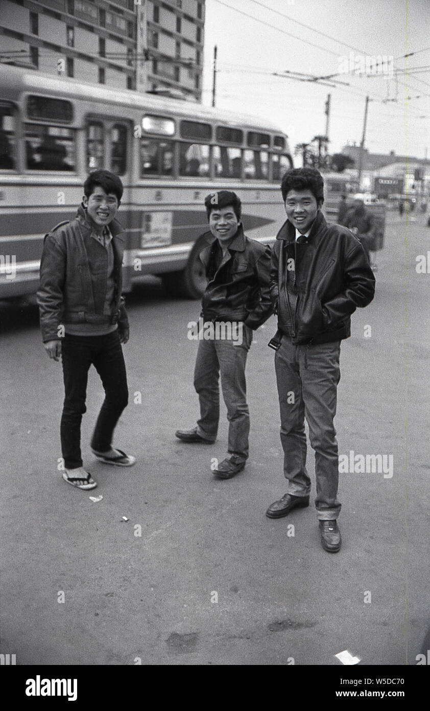 [1960s Japan - Trendige japanische Jugendliche] - Drei junge japanische Männer tragen trendige Frisuren, Jeans und Lederjacken, in der die Nachahmung des Amerikanischen Schmierer, stand auf einer Straße in Tokio als Stadt bus fährt, 1960 (Showa 35). Stockfoto