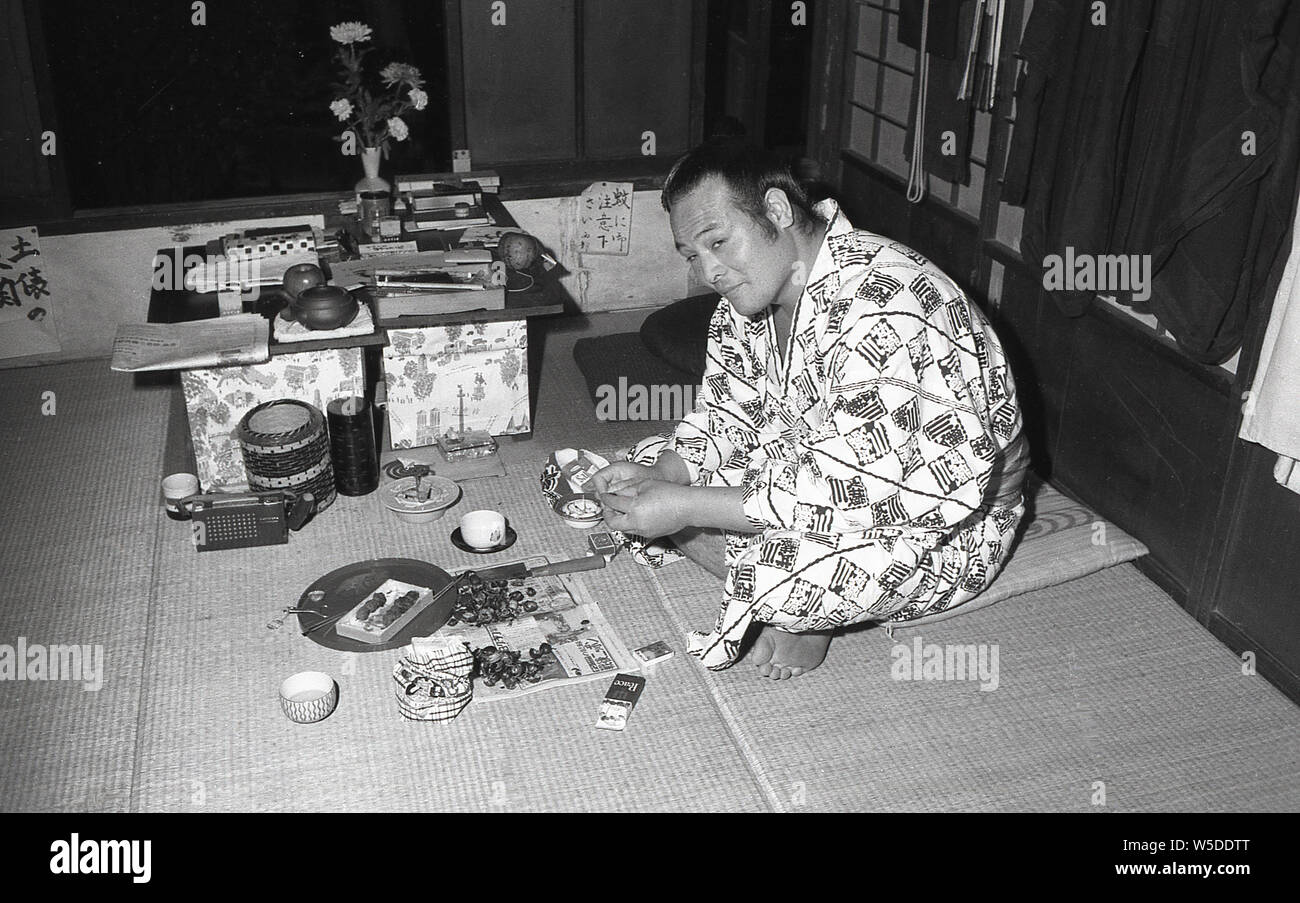 [1960s Japan - Japanische Sumo-ringer Ruhend] - ein Sumo-ringer im Yukata genießt einen Snack an Sojiji Tempel (総持寺) in Yokohama, Kanagawa Präfektur, 1960 (Showa 35). Ein kleines Transistorradio steht neben der Tabelle. Stockfoto