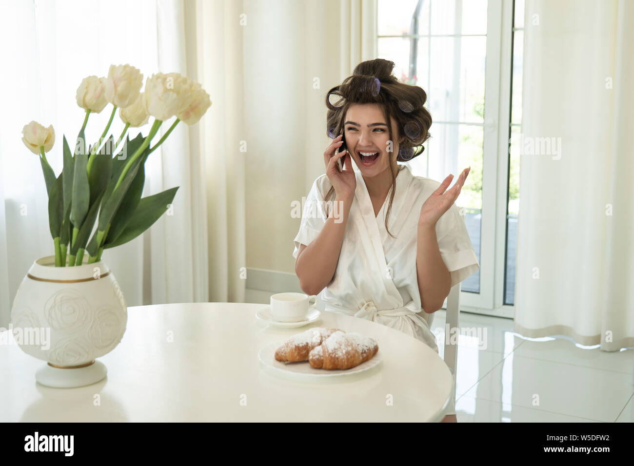 Schöne junge lächelnde brünette Frau in Lockenstäbe sprechen auf dem Smartphone beim trinken Kaffee und essen Croissants zum Frühstück im hellen Stockfoto