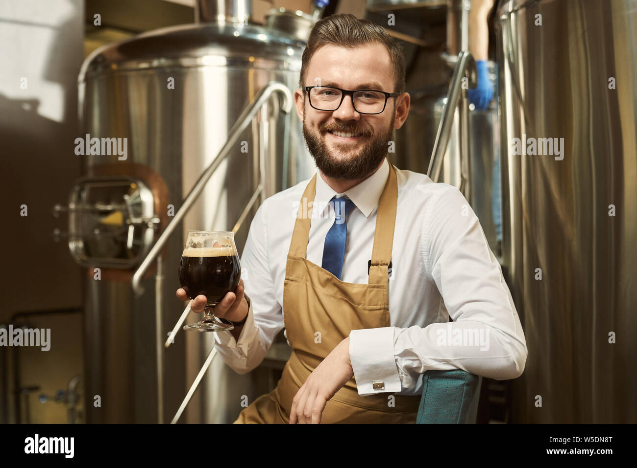 Positive, freundliche Brauer sitzen auf Schritte von Metall Plattform, Glas mit dunklem Bier. Stattliche Arbeiter Lächeln und Posieren auf dem Hintergrund von Stahl Behälter. Stockfoto