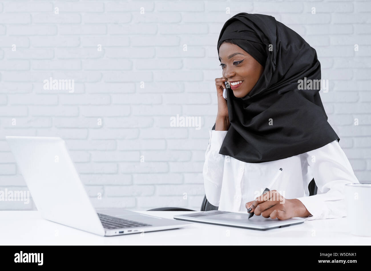 Ziemlich muslimische Frau Gespräch per Telefon, am Laptop suchen, lächelnd. Charmant, positive afrikanische Büro tragen in Schwarz hijab und weiße Kleidung am Tisch sitzen mit Computer. Stockfoto