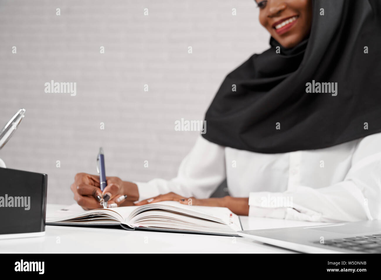 Close Up, 7/8 der jungen afrikanischen muslimische Frau am Arbeitsplatz und Schreiben im Notebook sitzen. Weibliche Büroangestellte in Schwarz hijab und weiße Kleidung holding Pen, lächelnd. Stockfoto